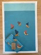 The Together Wild Swimming Tea Towel, a blue cotton kitchen accessory, features minimalist illustrations of six swimmers in swim caps. Displayed on wood, it comes with a folded version and tag on top.