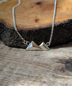 The Geometric Little Arran Necklace, featuring two small mountain-shaped pendants in sterling silver, displays its modern style against a rustic wood slice background.