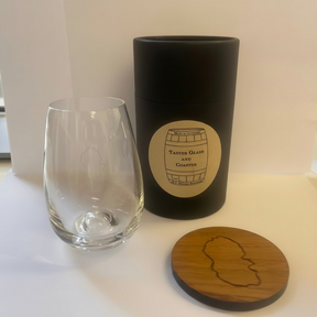 The Arran Whisky Shot Glass & Coaster Gift Set includes a clear whisky shot glass, a black cylindrical container, and a round wooden coaster with matching logos—a perfect Scotland souvenir displayed on a white background.