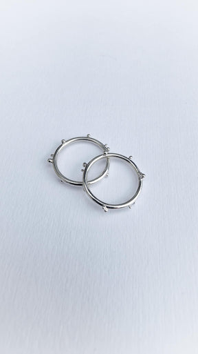 Two silver hoop earrings, adorned with small, evenly spaced protruding beads, rest on a textured white surface and gently overlap—showcasing a design inspired by the delicate elegance of the Dewdrop Ring collection.