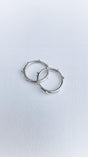 Two silver hoop earrings, adorned with small, evenly spaced protruding beads, rest on a textured white surface and gently overlap—showcasing a design inspired by the delicate elegance of the Dewdrop Ring collection.