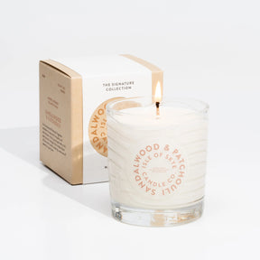 A lit Isle of Skye Sandalwood and Patchouli Tumbler Candle made from natural soya wax sits in a clear glass jar with its branded box featuring product details in the background.