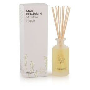The Max Benjamin – Meadow Hygge Reed Diffuser features natural essential oils in a frosted glass bottle with several reed sticks, hand-crafted in Ireland, and packaged in a white box decorated with green leaf illustrations.