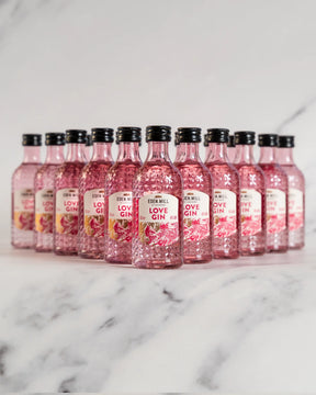 Twelve Eden Mill Love Gin Miniature 5cl bottles, small and clear pink with black caps and white labels featuring red and pink accents, are arranged in rows on a white marble surface against a light background.