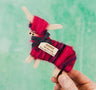 A person holds a Hairy Coo Standing Coo figurine, handcrafted with red and purple yarn, finished with a tartan ribbon and "ROWAN The Hairy Coo Handmade in Scotland" tag—ideal Scottish décor set against a green background.