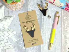 The Scottish Stag Wooden Brooch — All That Glitters, shaped like a stag’s head with antlers, sits on a brown tag on wood, surrounded by another brooch, pen, coin, potted plant, and floral fabric—a unique Scottish heritage gift.