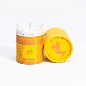 The Isle of Skye Highland Gorse Miniature Candle, a handmade white soya wax candle in a glass jar with a yellow and orange plaid label, sits by its matching cylinder container lying on its side.
