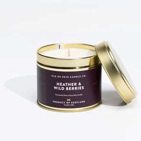 An Isle of Skye Heather and Wild Berries Travel Tin Candle by Isle of Skye Candle Co. is displayed with its lid resting nearby. The white candle comes in a gold tin, featuring a label that describes it as a handmade natural soya wax candle from the Isle of Skye, infused with Scottish heather harvested in Scotland.