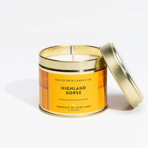 A gold tin labeled “ISLE OF SKYE CANDLE CO. HIGHLAND GORSE” sits partially open, showing a white candle inside. The handmade, natural soya wax candle is the Isle of Skye Highland Gorse Travel Tin.