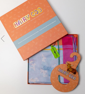 The Hairy Coo Scarf comes in an orange "Hairy Coo" box with cute cow illustrations and a cow-shaped tag. Made from recycled fabric, it features blue and pink plaid with a green "re:preve" label—a perfect Scottish gift.
