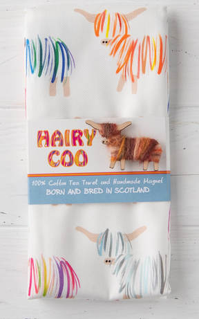 The Hairy Coo Tea Towel is a white cotton towel with colorful Highland cows, hand-wrapped and paired with a cute coo magnet. The label says "HAIRY COO" and "Born and Bred in Scotland.