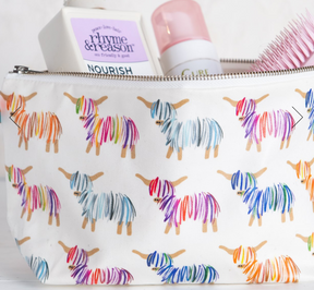 The Hairy Coo Zip Bag is a white cotton bag featuring colorful Hairy Coo illustrations, shown partially open with hair products and a pink hairbrush inside.