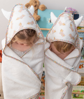 Two young children stand side by side, wrapped in Baby Coo Hooded Towels with colorful trim and small horns, both looking down as shelves filled with books and toys fill the background.