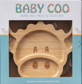 The boxed Baby Coo – Highland Cow Bamboo Plate is an eco-friendly baby plate shaped like a Highland cow face, with eye, nose, and ear cutouts. The packaging says "Born and bred in Scotland" and "Natural bamboo suction plate.