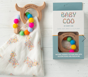 The Baby Coo - Highland Cow Teething Set, featuring a wooden teething ring with colorful silicone beads and a white muslin bib with highland cow prints, is displayed next to its packaging on a white wooden background.
