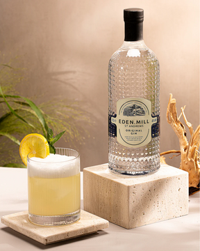 A bottle of Eden Mill Original Gin 70cl is displayed on a stone block beside a cocktail garnished with lemon and fresh herbs, both resting on light stone surfaces against a soft, natural backdrop.