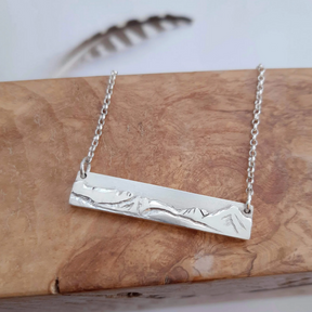 A sterling silver necklace featuring a rectangular pendant engraved with a mountain range, displayed on a wooden surface. This handcrafted jewellery piece, known as the Isle of Arran Skyline Necklace, is beautifully complemented by a blurred feather in the background.