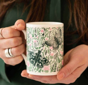 A person holds the Cherith Harrison Thistle & Butterfly China Mug, a hand-decorated UK mug adorned with green leaves, pink flowers, and two butterflies. Their nails are unpainted, and they wear silver rings on their fingers.