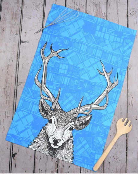 The Tartan Stag Tea Towel by Gillian Kyle features a black and white illustration of a deer’s head with large antlers on blue fabric, displayed on a wooden surface next to a whisk and wooden salad fork.