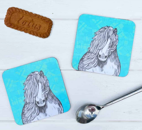 The Tartan Pony Set of 2 Coasters features blue coasters with black and white illustrations of a Shetland pony, shown with a silver spoon and Lotus biscuit on a white wooden surface.
