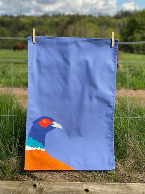 The Pheasant Tea Towel hangs on a clothesline outdoors, its vibrant purple pheasant design bringing charm to a country kitchen scene with green grass, a fence, and trees beneath a partly cloudy sky.