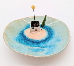 The Ceramic House Dish features a blue, water-like center, a small white house with a brown roof, yellow ball on wire, and green tree—evoking a tiny island scene and making it an ideal handmade gift.