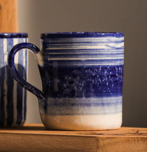A Selbrae House Ripple Ceramic Mug, hand-painted with blue and white stripes, sits on a wooden shelf. It features a curved handle, slightly textured surface, and another mug is partially visible in the background.