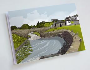Illustration of a stone bridge over a flowing river, with green grass and a stone path. Houses and trees under a cloudy sky—featured on the Turadh Design Blackwaterfoot Card, perfect for Isle of Arran-themed stationery.