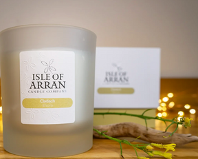 A frosted Cladach Shore Candle sits on a wooden surface beside a yellow flower, driftwood, and fairy lights, evoking an uplifting Arran sea breeze, with its blurred branded box visible in the background.