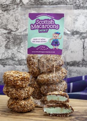 A clear bag labeled "Mint Scottish Macaroons" from Kilted Fudge Company sits on a wooden surface, filled with coconut-covered macaroons and one split to show its pale green mint filling. A tartan cloth is visible in the background.