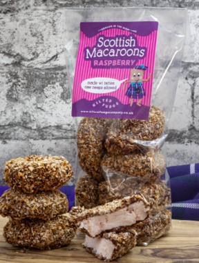 A standing bag of Raspberry Scottish Macaroons displays coconut-covered treats, some split to show their pink center. Set against a stone wall and tartan cloth, the scene reflects the charm of Scottish Highlands fudge.