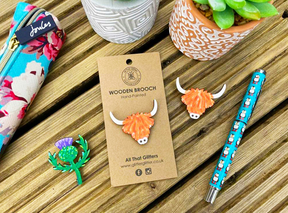 The Highland Coo Wooden Brooch, thistle pin, floral pencil case, cow-patterned pen, and two potted plants are displayed on wood—making a unique Scottish-themed gift.