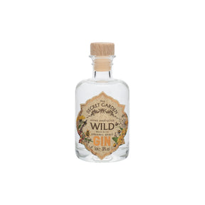 A 5cl clear glass bottle of Secret Garden Distillery Wild Gin Miniature with a wooden cork, featuring an ornate label with botanical illustrations stating it is hand-harvested and organic, is pictured on a white background.