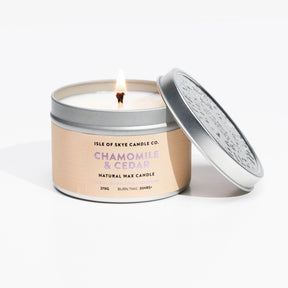 A lit Chamomile & Cedar Candle Travel Tin from Isle of Skye Candle Company sits with the lid partly removed, its flame visible. The round metal tin offers a calming chamomile and cedar fragrance.