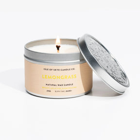 A handmade Lemongrass Travel Tin candle by Isle of Skye Candle Company sits lit, with its decorative lid beside it. Its label shows: “Natural Wax Candle, 270g, Burn time: 35hrs+.” The round tin exudes a fresh lemongrass scent.