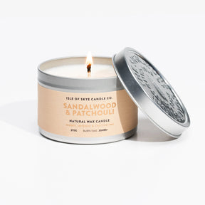 A lit Isle of Skye Sandalwood and Patchouli Candle Travel Tin labeled “Sandalwood & Patchouli” with the lid beside it. The tan label shows “Isle of Skye Candle Co.,” handmade in Scotland, plus scent info and burn time.