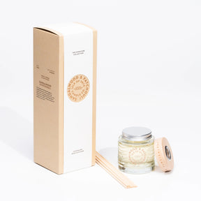 A clear glass jar of Isle of Skye Candle Company Sandalwood and Patchouli Reed Diffuser with a wooden lid, reed sticks, and a tall beige box, all set against a white background.