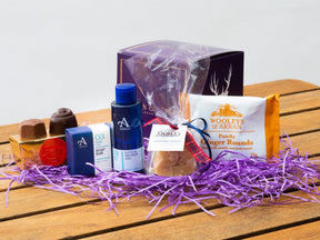 The Wee Sannox Arran Gift Box includes Scottish skincare from the Isle of Arran, chocolate, blue bath and shower gel, a soap bar, shortbread, and ginger rounds in a purple box on shredded paper.