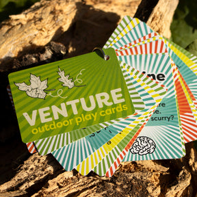 A set of colorful VENTURE Outdoor Play Cards for nature exploration, fanned out on tree bark. The green front card shows the product name and two leaf drawings—ideal outdoor activity cards for kids.