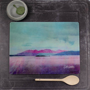 The Arran Glass Worktop Saver, decorated with pastel Scottish landscapes of mountains and water, rests on a dark surface. Nearby are a wooden spoon and a small bowl with a green leaf.