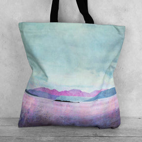 The Tote Bag Isle of Arran features black handles and a pastel landscape of mountains and lakes in blue, purple, and pink on a light background. Ideal for those who love spacious tote bags with distinctive designs.