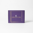 A large Arran Gift Box in purple, featuring "Isle of Arran, Scotland" in gold lettering, rests on a rock by a lakeside. Majestic mountains and a partly cloudy sky grace the background, creating an ideal setting for savoring exquisite Scottish delicacies.