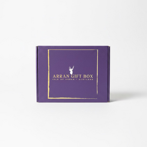 A large Arran Gift Box in purple, featuring "Isle of Arran, Scotland" in gold lettering, rests on a rock by a lakeside. Majestic mountains and a partly cloudy sky grace the background, creating an ideal setting for savoring exquisite Scottish delicacies.