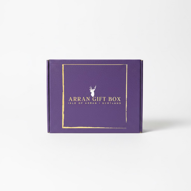 A large Arran Gift Box in purple, featuring "Isle of Arran, Scotland" in gold lettering, rests on a rock by a lakeside. Majestic mountains and a partly cloudy sky grace the background, creating an ideal setting for savoring exquisite Scottish delicacies.