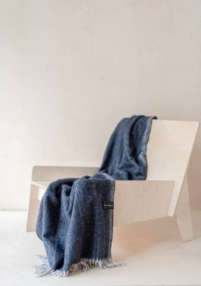 A minimalist light wood chair rests against a pale backdrop, draped with the Recycled Wool Knee Blanket in Navy Herringbone. The eco-conscious blanket features subtle patterns and white fringe, adding sustainable style to the serene setting.