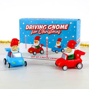 Two toy gnomes in blue and red race cars stand in front of the Driving Gnome For Christmas box, which showcases festive snowy graphics and cheerful images of gnomes racing. This playful set makes a charming stocking stuffer that brings extra holiday spirit to your celebrations.