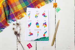 The Scottish Isles Watercolour Map Greetings Card, featuring vibrant island maps with labeled names, is displayed on a white wooden surface alongside tartan fabric, a gold pen, binder clip, dried flowers, and postage stamps.