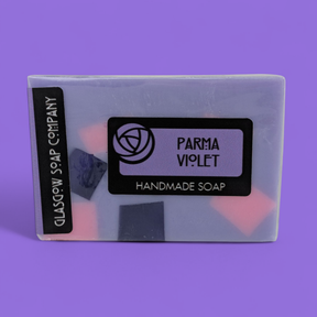 A rectangular Parma Violet Handmade Soap Slice with a light purple hue and embedded pink and dark purple pieces. The label displays "Glasgow Soap Company," "Parma Violet Soap," and "Handmade Soap" on a purple background.