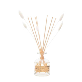 The Atacama Reed Diffuser White comes in a clear glass bottle with a brown label, featuring sustainably sourced reeds and white-tipped sticks to spread its citrus and wood fragrance, elegantly showcased against a plain white background.