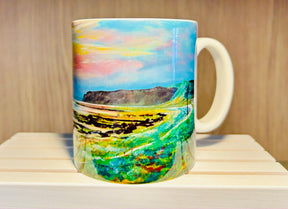 The Bennan Head Mug is a white ceramic mug featuring a vibrant landscape painting of grassy fields, winding paths, distant hills, and a bright sky with clouds, displayed on a light wooden shelf.